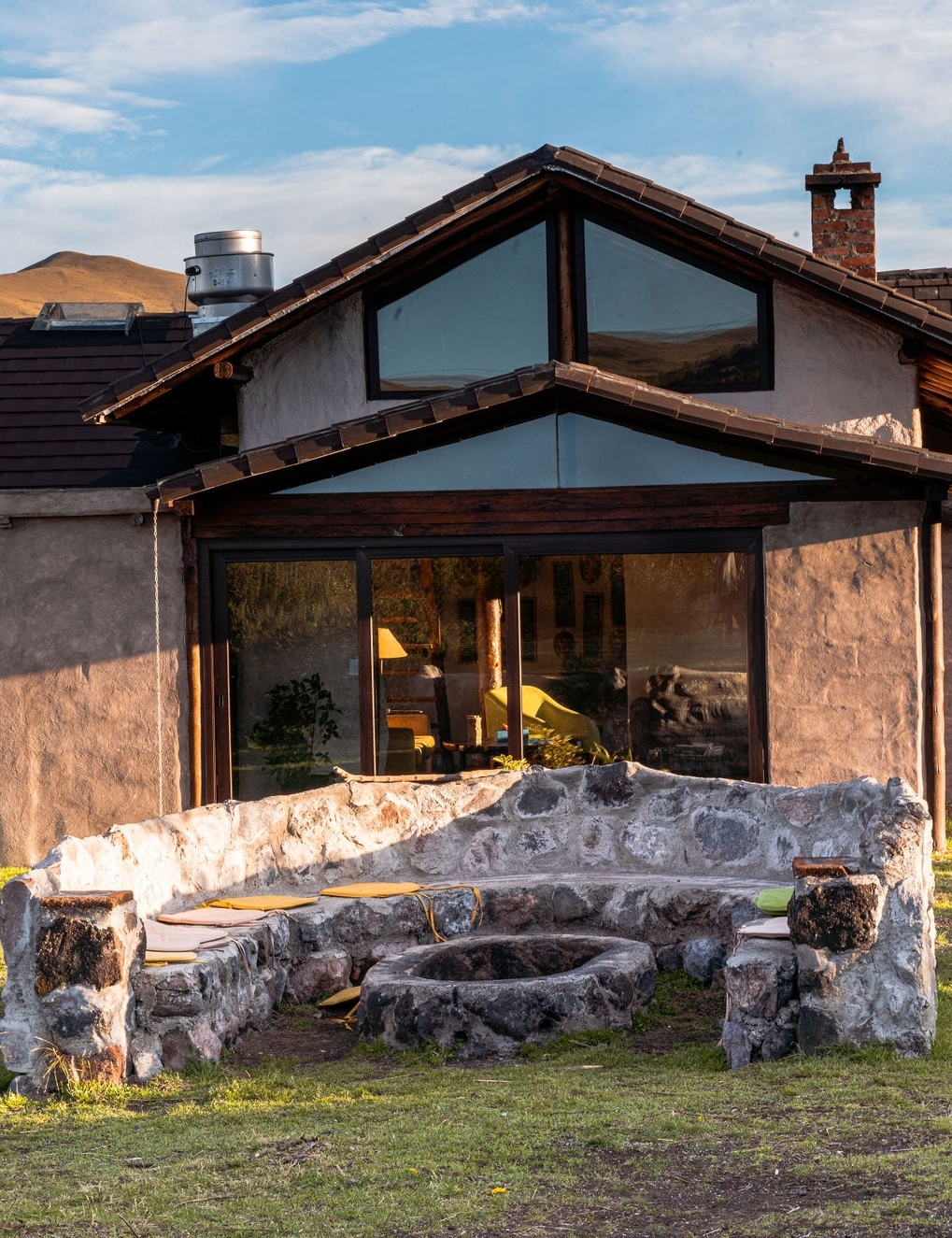 A rustic cabin with large windows and a stone-built outdoor bench in a grassy area.