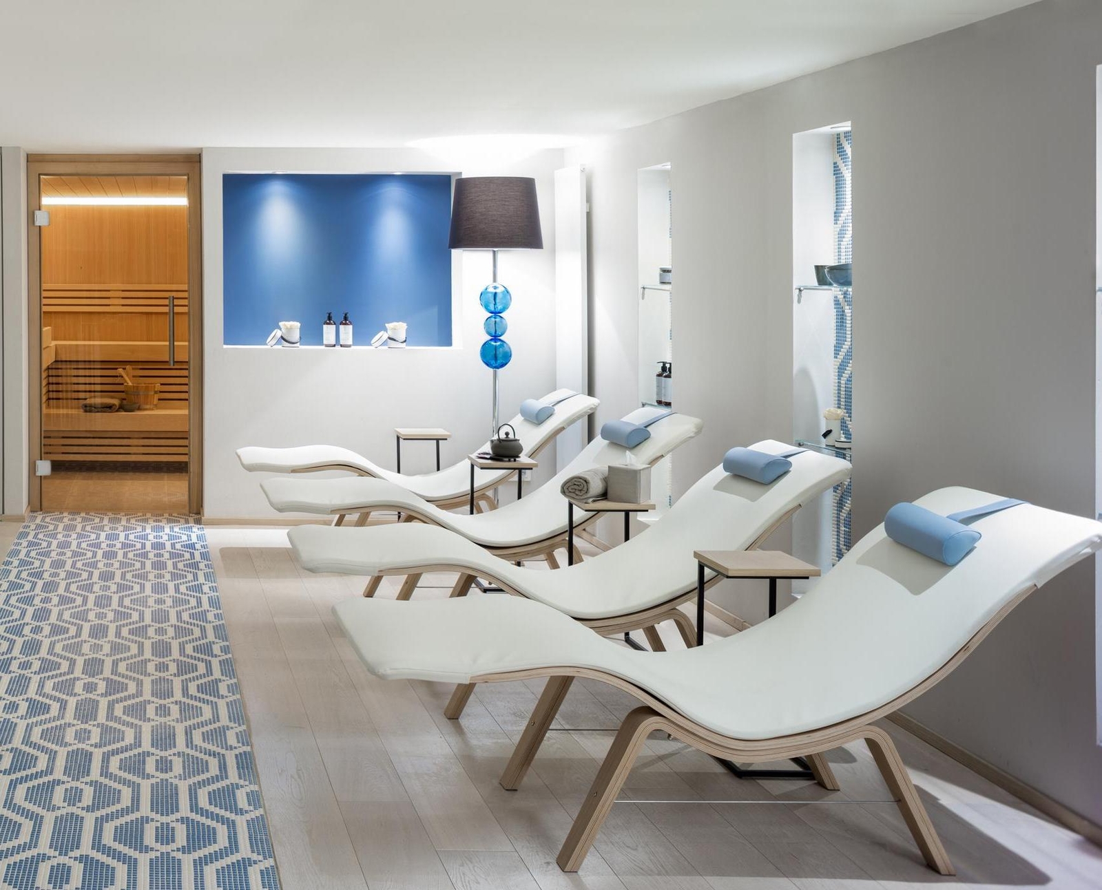 Modern spa relaxation room with white loungers, blue accents, and an open sauna door.