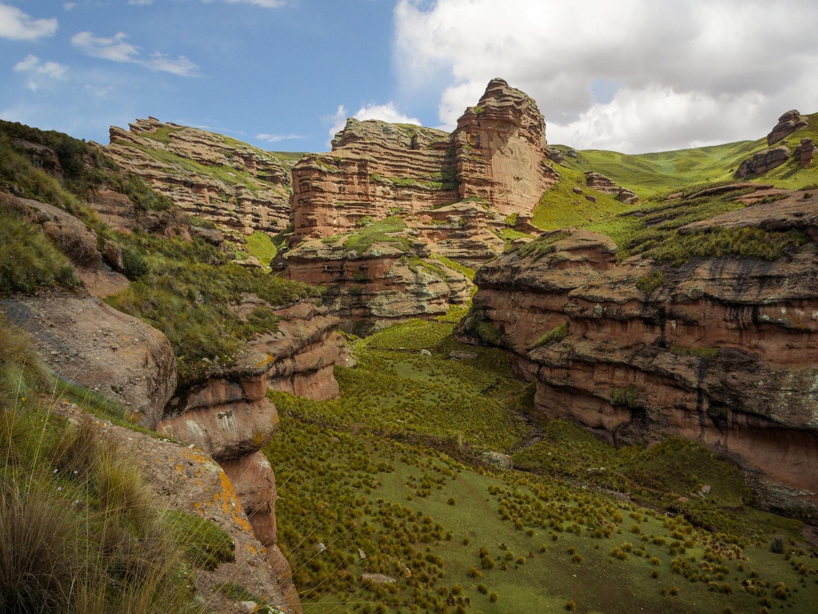 Layered sandstone cliffs with greenery in a mountainous landscape under a cloudy sky.