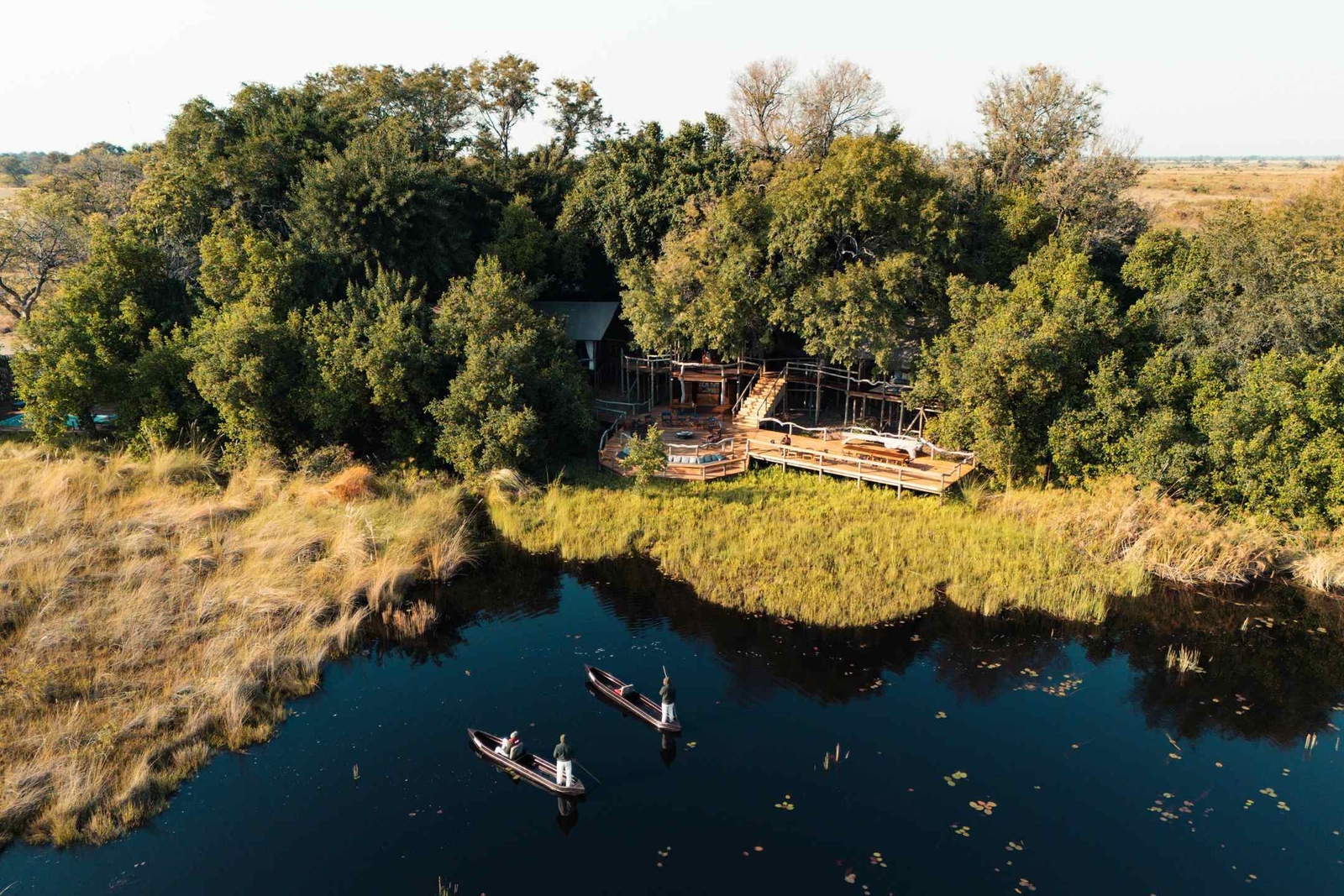 Aerial of Shinde Camp which sits at the edge of a lagoon, where two men steer a pair of traditional canoes.