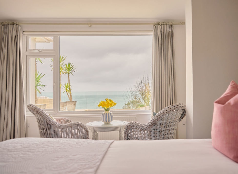 Hotel room with wicker chairs and yellow flowers facing a large window with ocean views at Boskerris Hotel Cornwall.