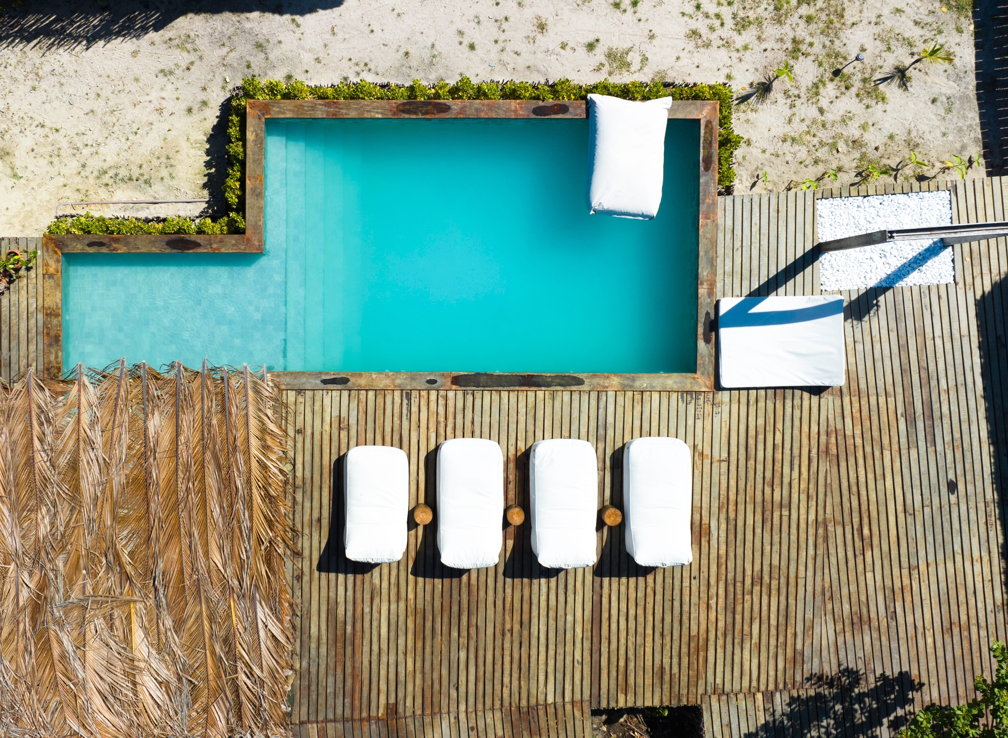 On overhead view of a swimming pool with blue water and four white deck chairs and some white towels next to it.