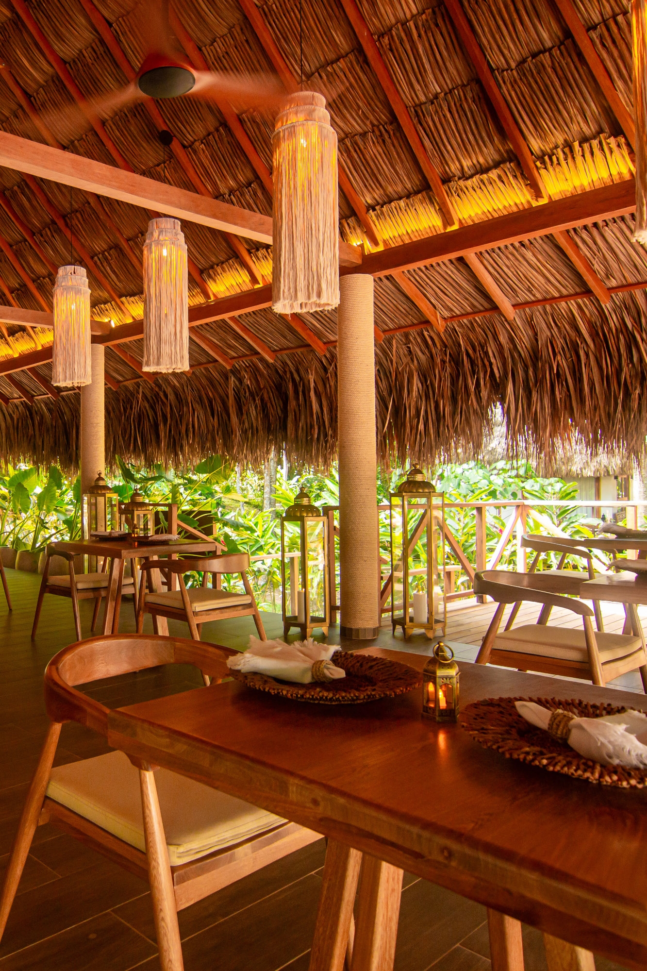 An open-air restaurant dining area with wooden tables and a low-hanging thatched roof. Inside are three long wooden lamp shades hanging from the roof. Outside you can see dense green foliage.