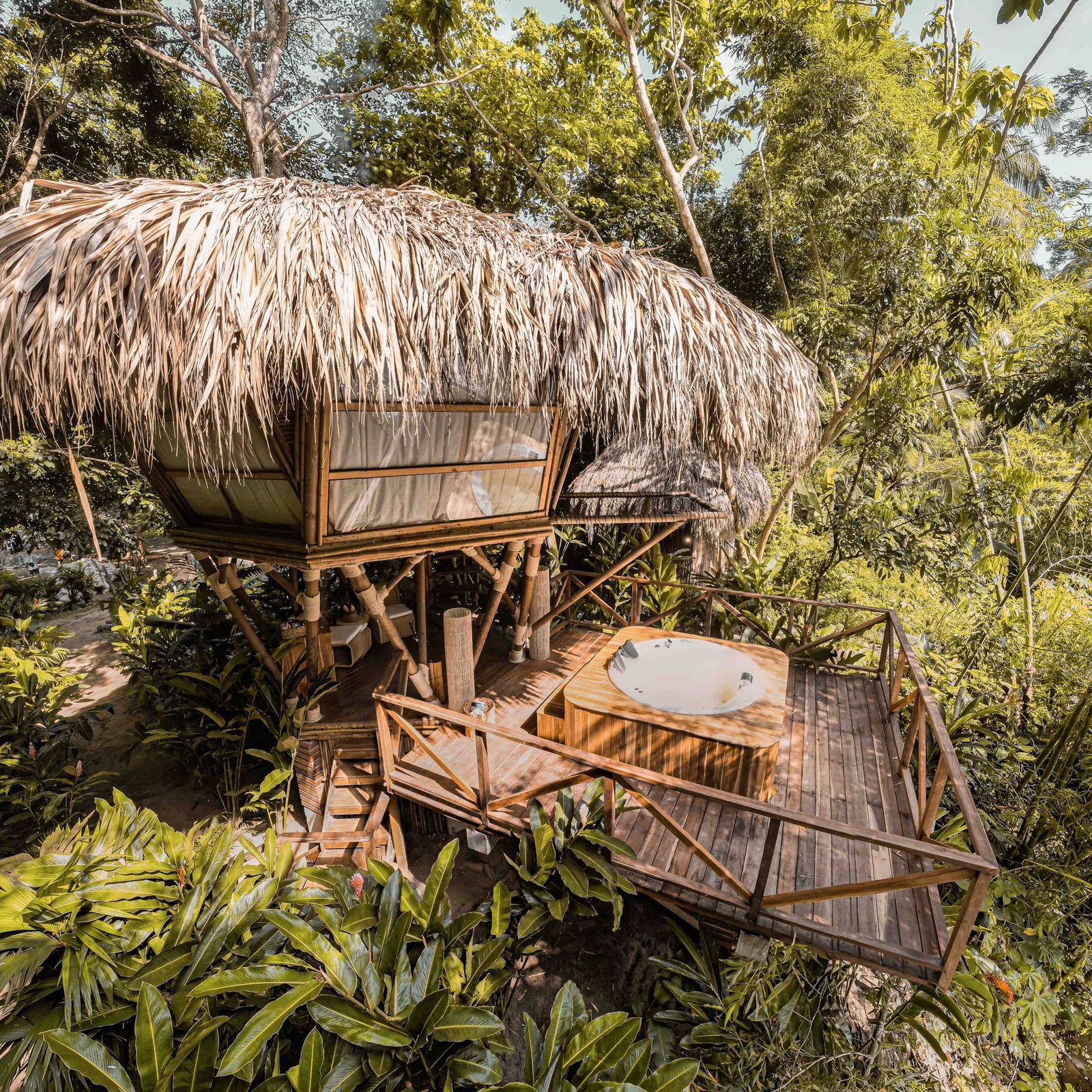 View of a wooden treehouse with long hanging thatched roof. It is surrounded by a wooden deck with a hot tub and in the background and foreground is dense green jungle foliage.