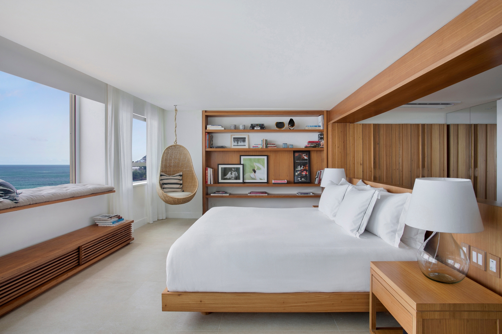 A bedroom with light wood bed and bedside table and shelves. The bed has white sheets and pillows, and there is a white lamp on the bedside table. The bed is facing a picture window on the left that looks out to a blue sea and sky. In the corner is a hanging basket swing chair.