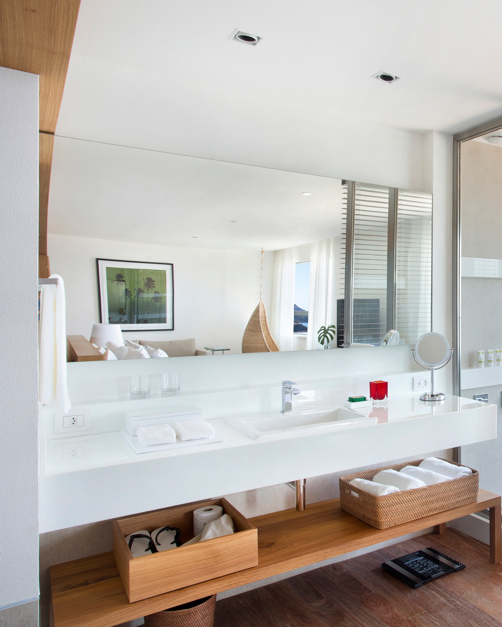 A bathroom vanity with sink and white counter underneath which are light wood shelves and drawers full of towels and toilet paper. Over the sink is a mirror depicting a bedroom with a framed green painting.