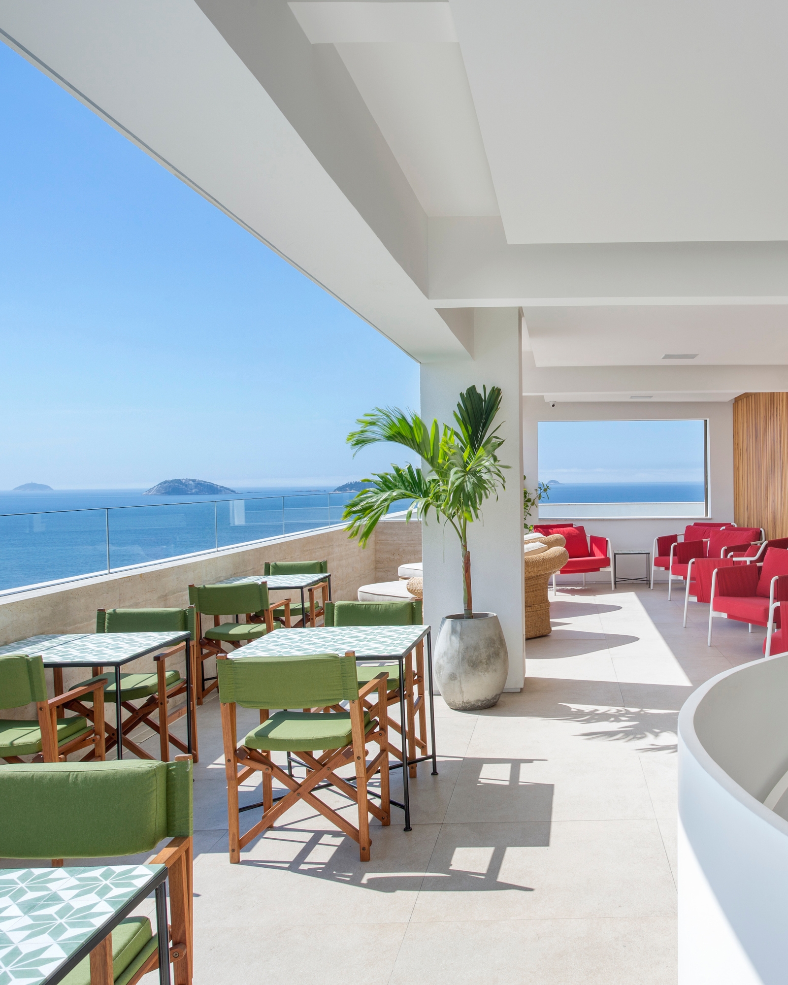 An open-air terrace with white ceiling and floor. On the right are red lounge chairs, on the left are green chairs and dining tables. In the background is a clear glass railing and beyond is the blue sea and sky with a few islands in the distance.