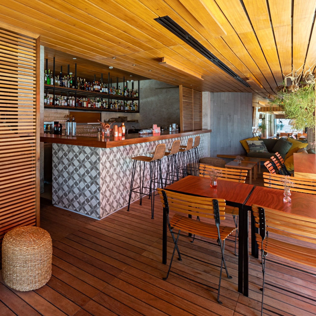 Entryway to a restaurant with a bar area on the left with tiled counter, wooden stools and open shelves above with bottles of alcohol. On the right are some wooden tables and chairs. The floor and ceiling are both made of hard wood.
