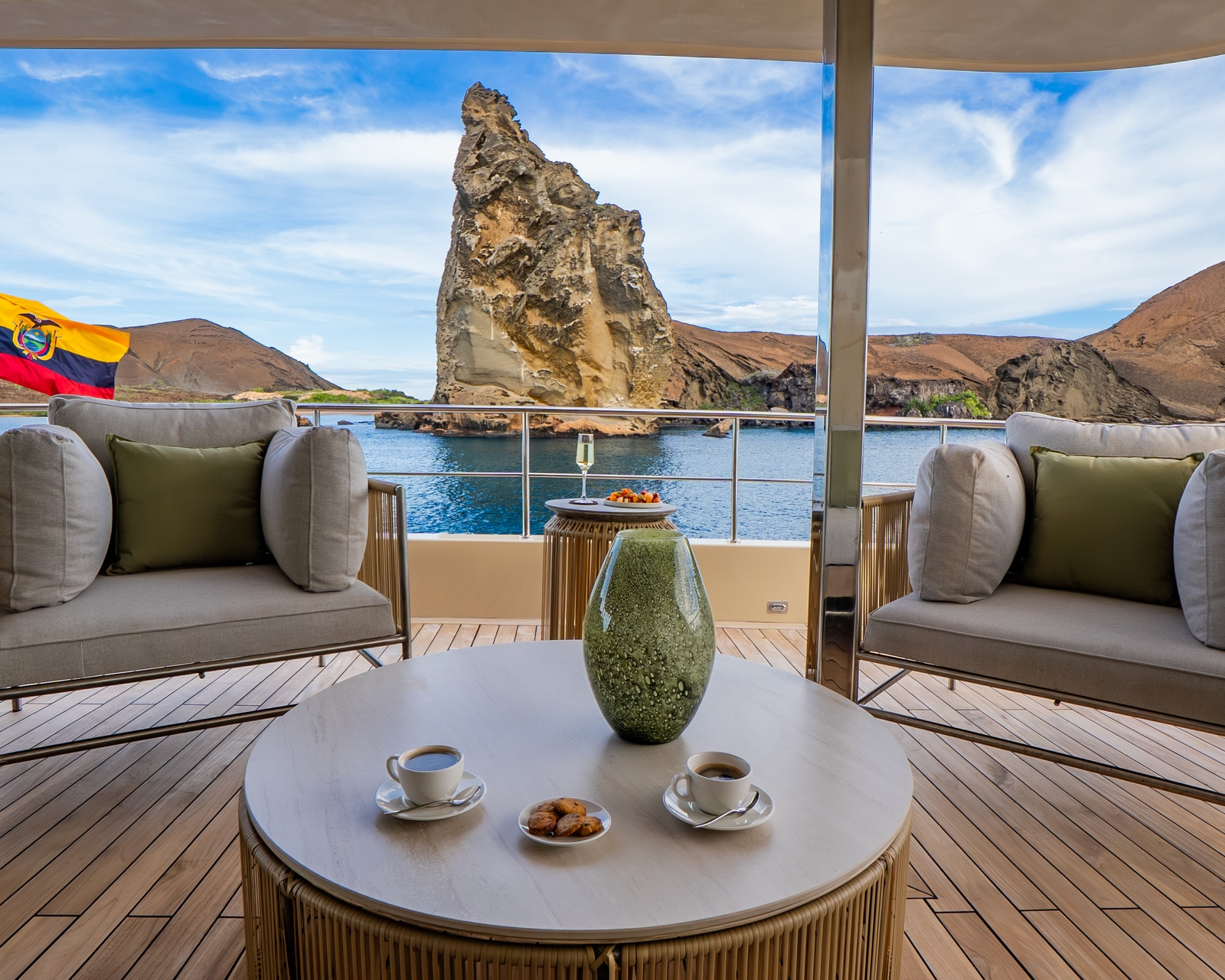 Plush outdoor armchairs and coffee tables with refreshments on a wooden deck overlook a towering volcanic rock island.