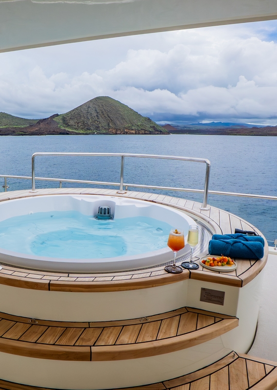 A circular hot tub filled with blue water on a wooden deck, with cocktails nearby and a volcanic island in the distance.