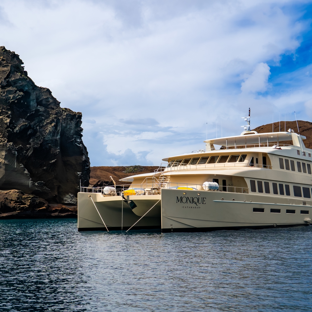 The Monique catamaran floats on calm blue ocean waters next to massive volcanic rock cliffs under a cloudy sky.