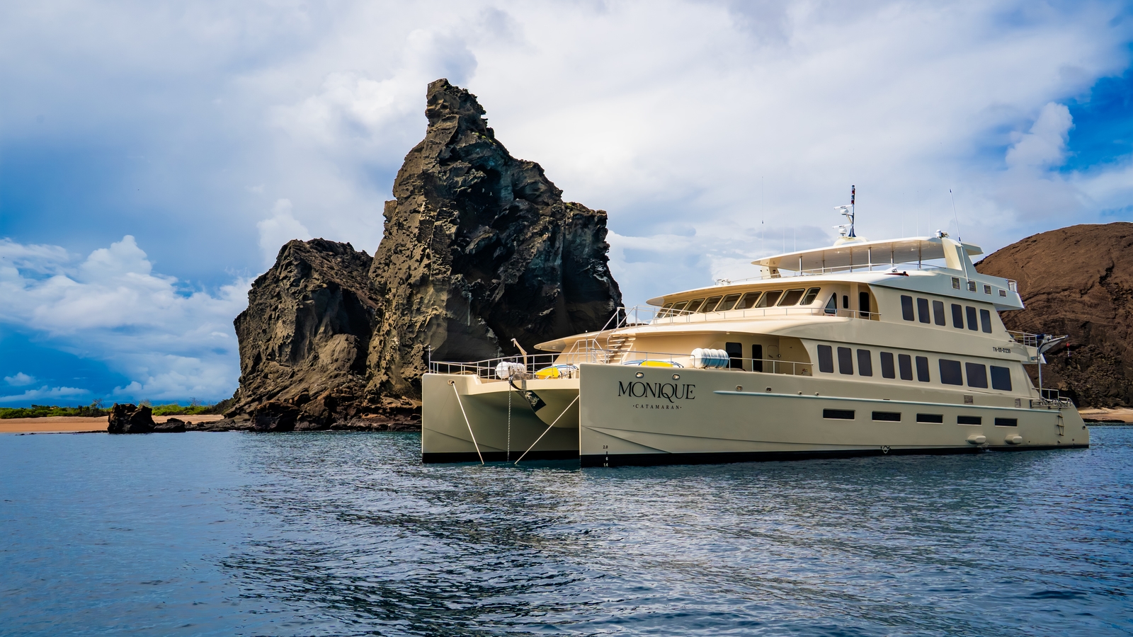 The Monique catamaran sails across the ocean past a towering, rugged volcanic rock island under a stormy sky.