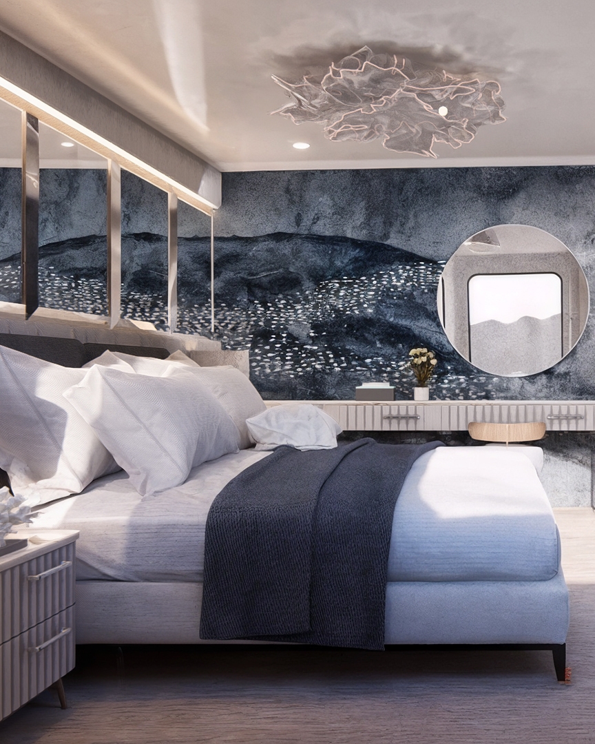 A bedroom with a double bed with grey and white sheets and pillows, a crystal chandelier, white carpeting and a round vanity mirror along a dark grey wall.