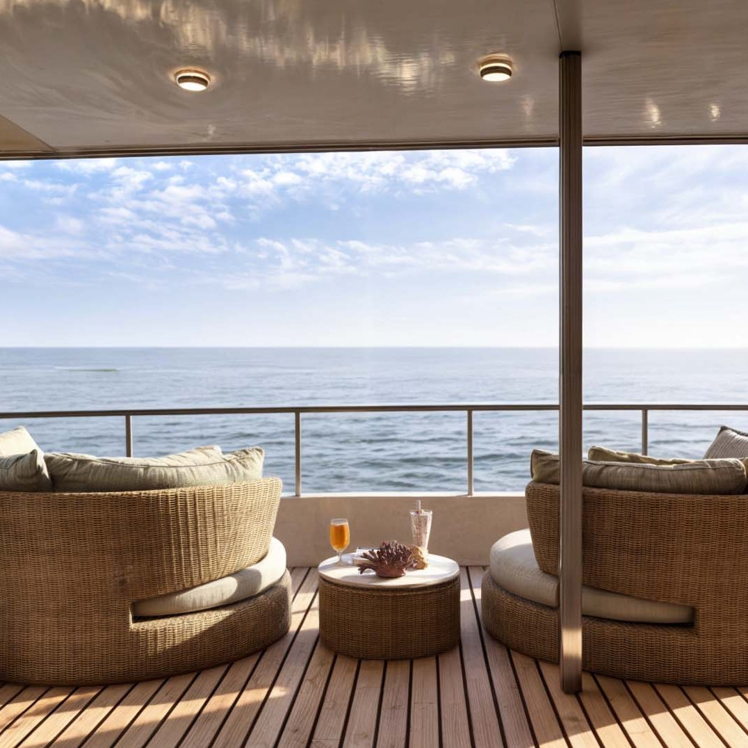 An open-air boat deck with a two rattan lounge chairs and small table between them with cocktails. In the background, a railing of the boat deck, and the sea and blue sky with white clouds.