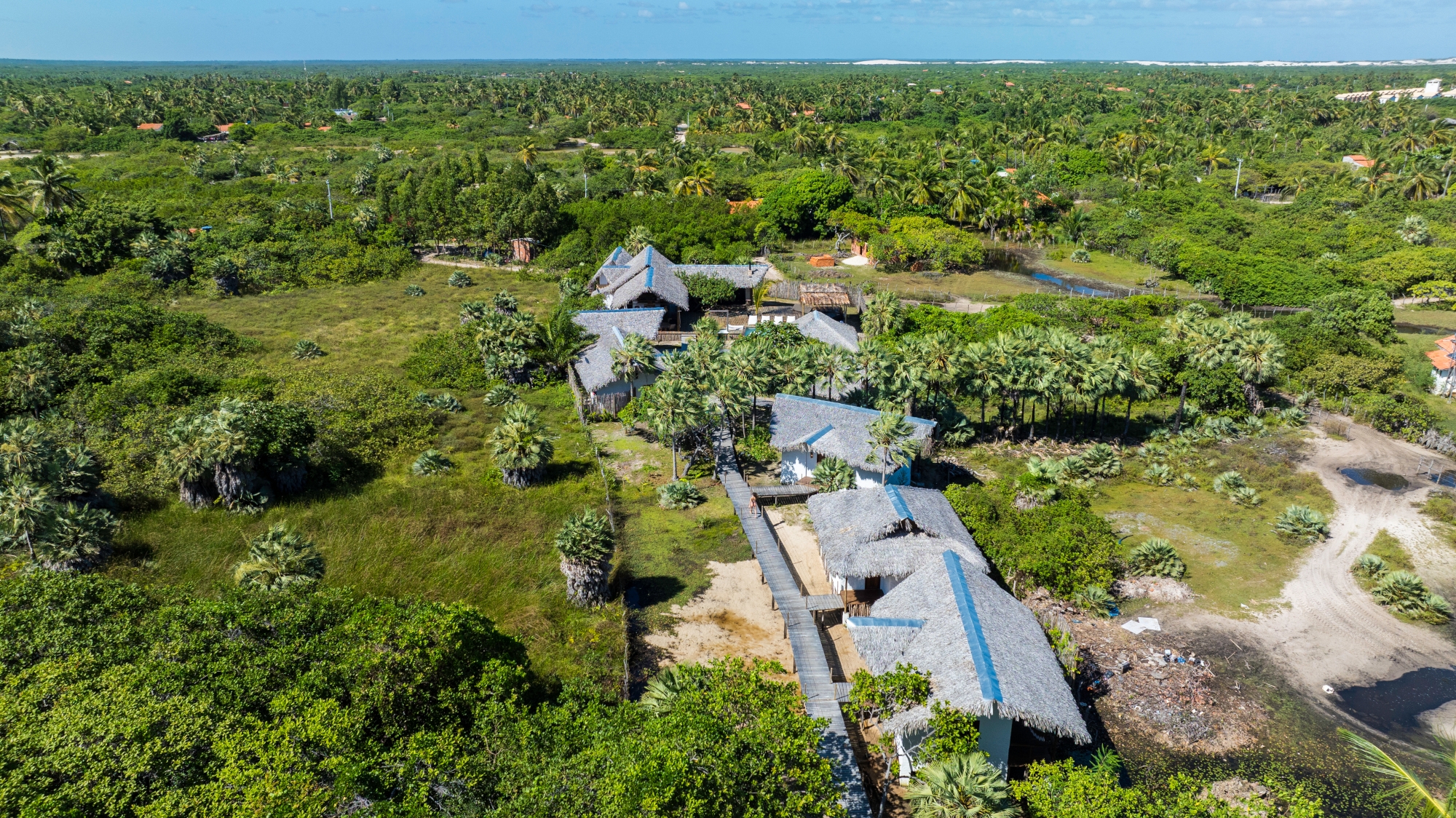 An overhead view of lodge buildings with thatched roofs set in amongst green jungle with a small dirt road on the right and blue sky above.