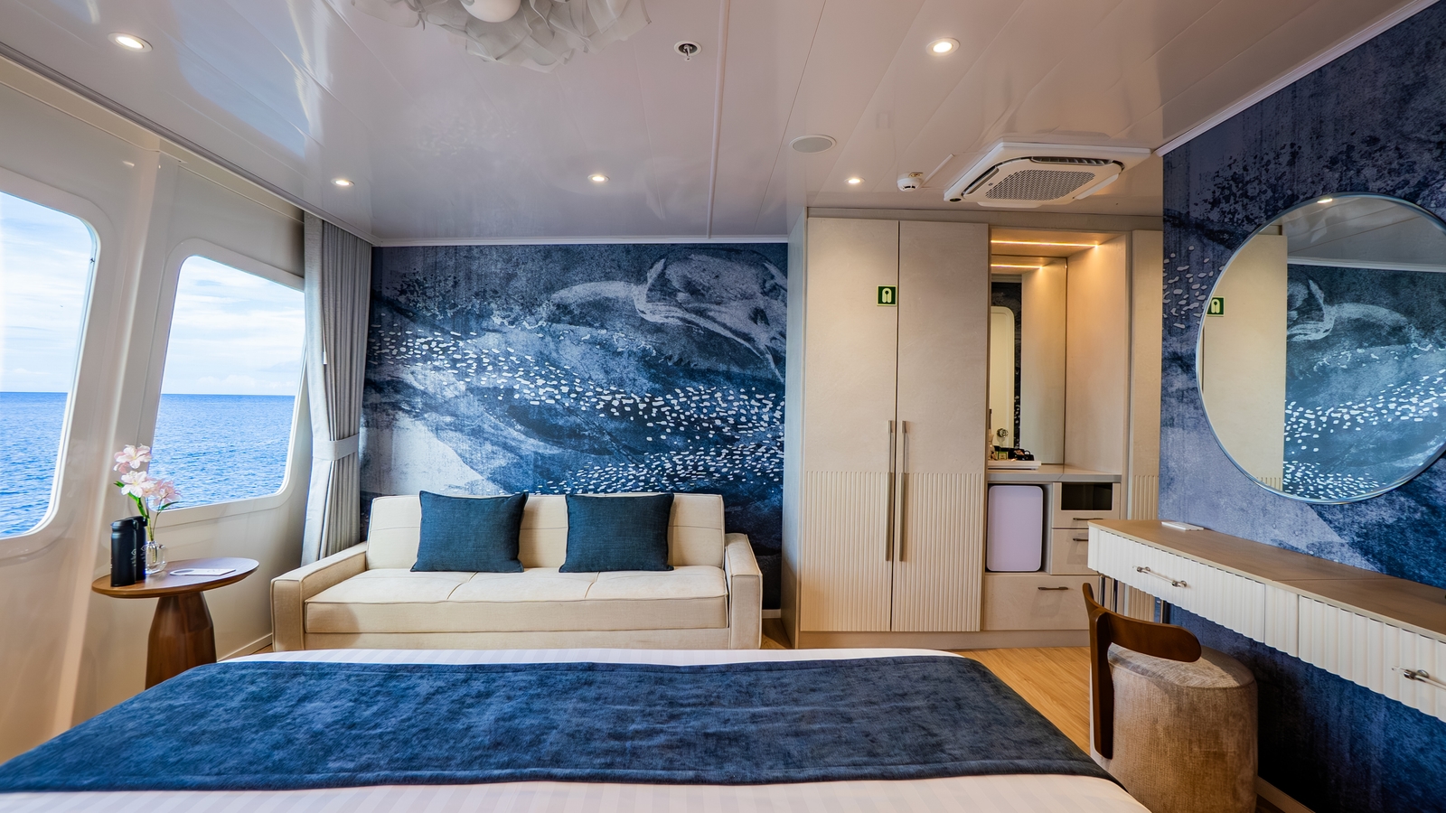 A luxury yacht cabin interior with a large bed, a cream-colored sofa, and a wide window displaying the blue ocean.