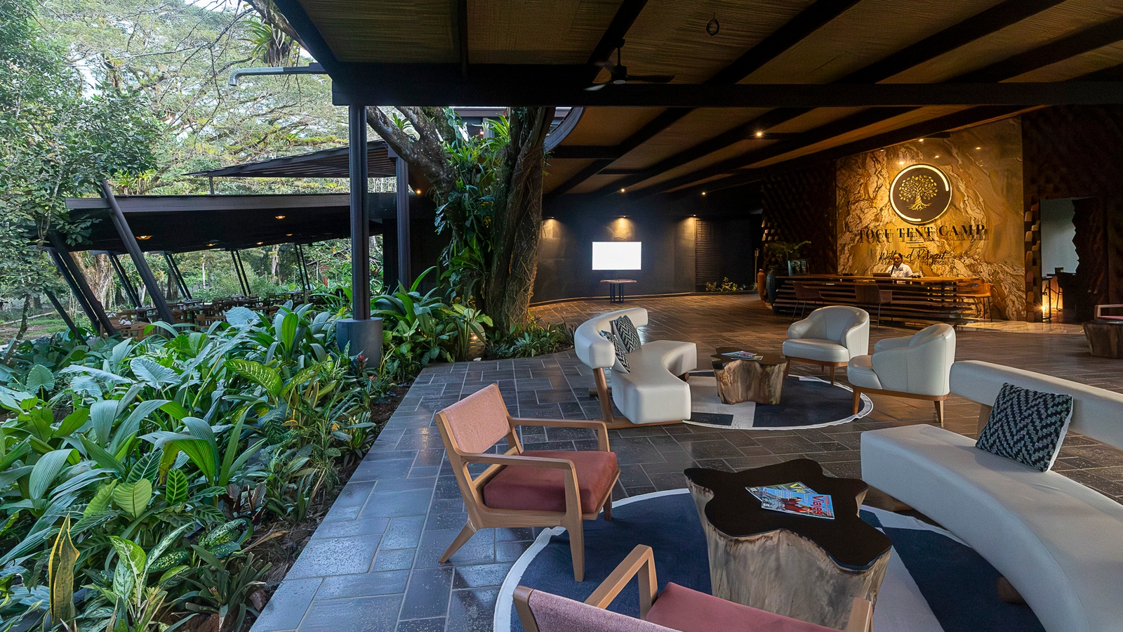 An open-air hotel reception area with a covered wooden deck where there are lounge chairs and sofas overlooking jungle greenery.