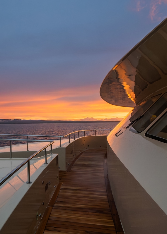 A glossy wooden walkway on a yacht deck reflects the vibrant orange and pink colors of a sunset over the sea.