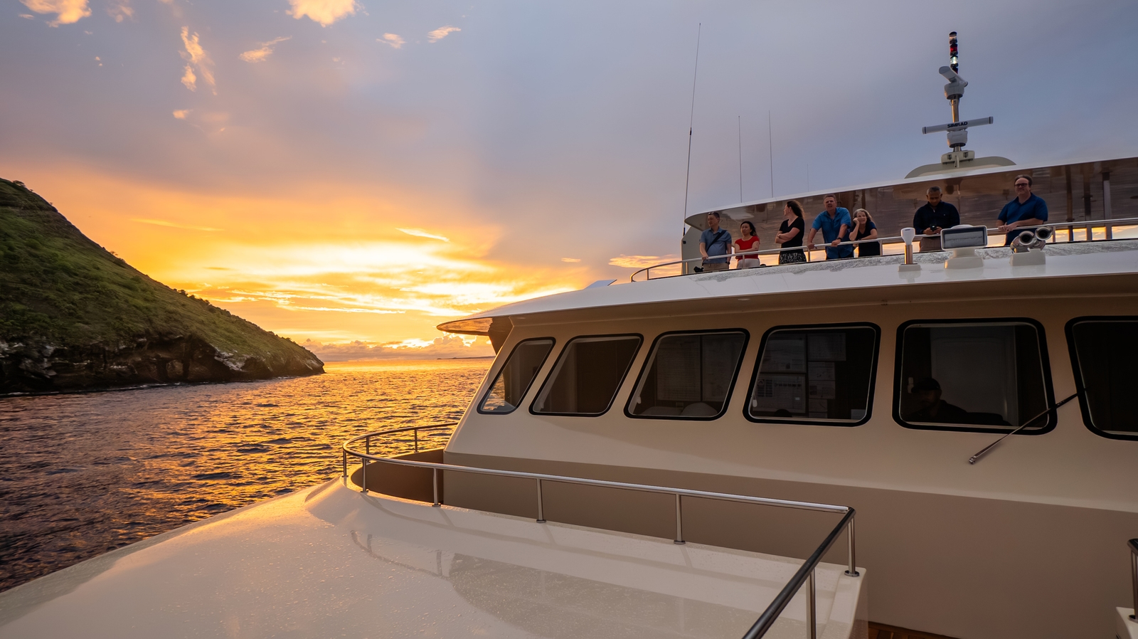 Passengers stand on a yacht upper deck looking out at a vibrant orange and yellow sunset over the water.