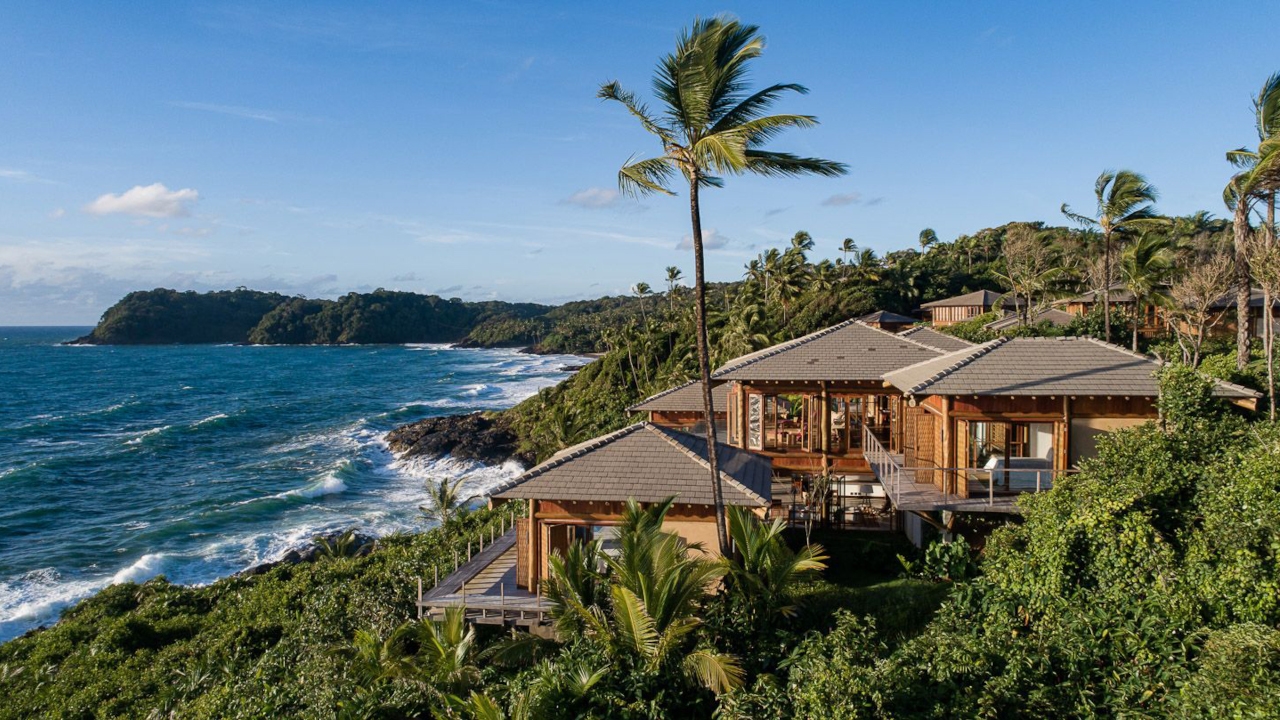 Villa with pitched roof and wrap-around terraces set in thick palm jungle overlooking a rocky coastline with blue ocean and waves crashing.