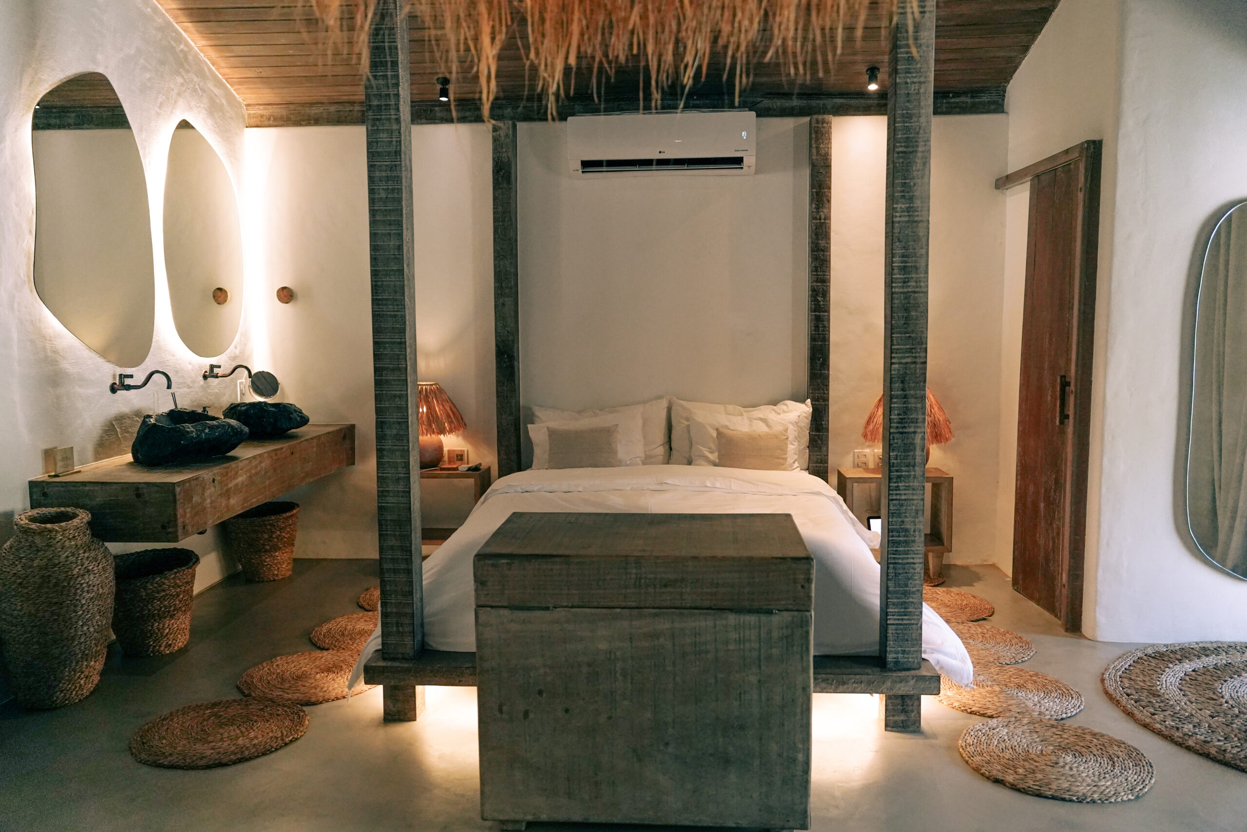 Interior of a bedroom – a double bed with white sheets and grey-wood four poster beams, a grey-wood cabinet at the foot of the bed, rugs on the floor, white walls and an odd-shaped mirror on the far wall with two sinks carved out of dark rocks.