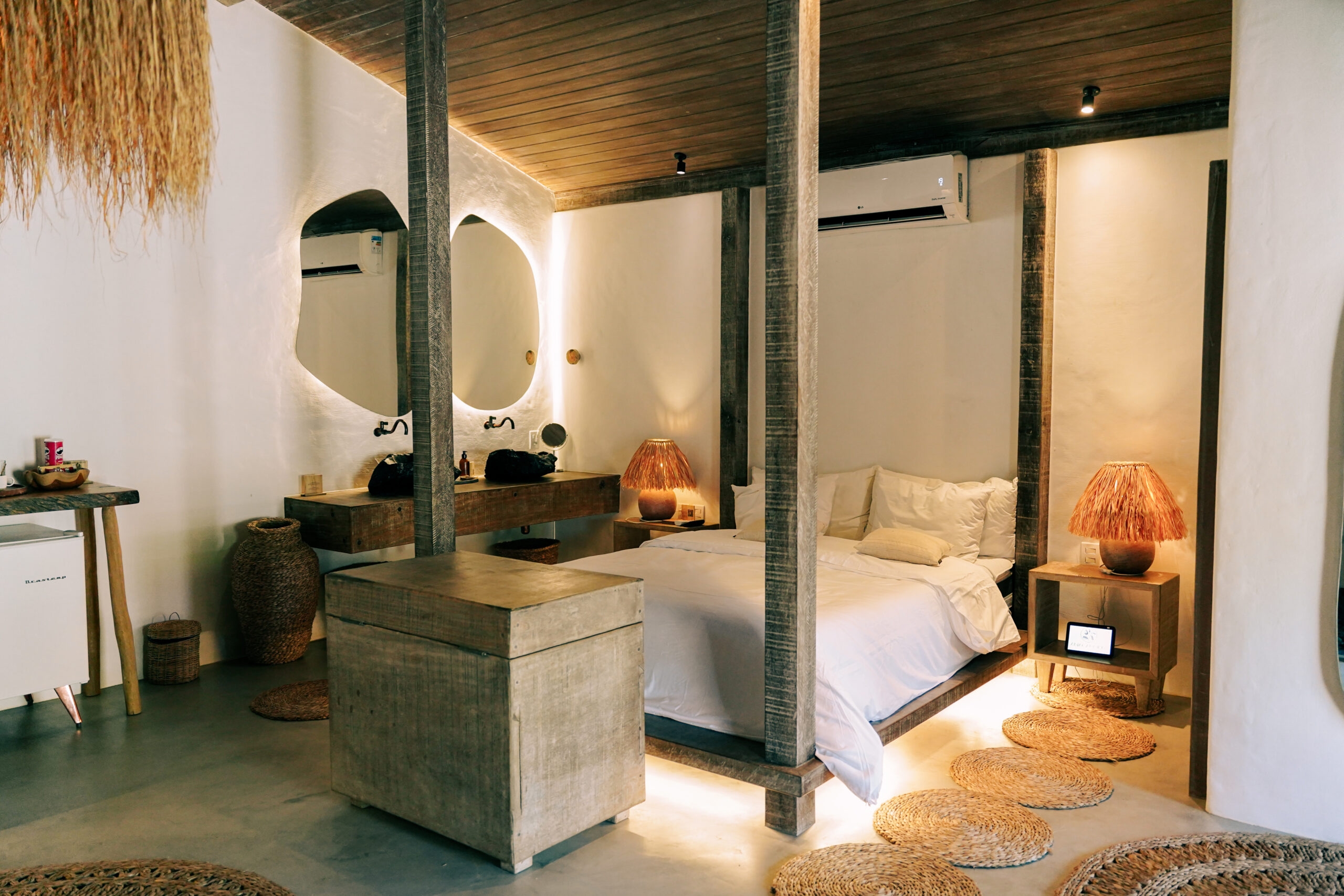 Interior of a bedroom – a double bed with white sheets and grey-wood four poster beams, a grey-wood cabinet at the foot of the bed, rugs on the floor, white walls and an odd-shaped mirror on the far wall with two sinks carved out of dark rocks.