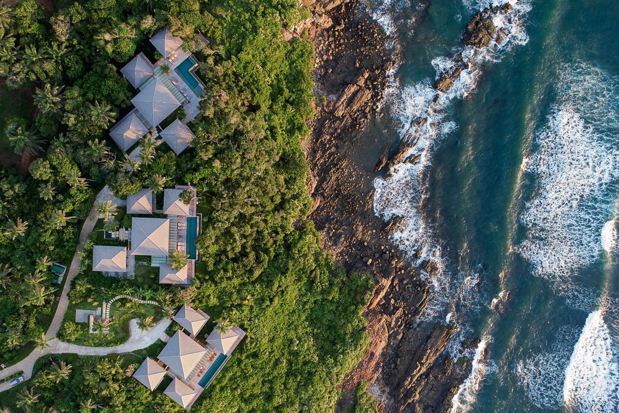 Aerial view of a village with multiple sections set in thick palm jungle overlooking a rocky coastline with blue ocean and waves crashing.