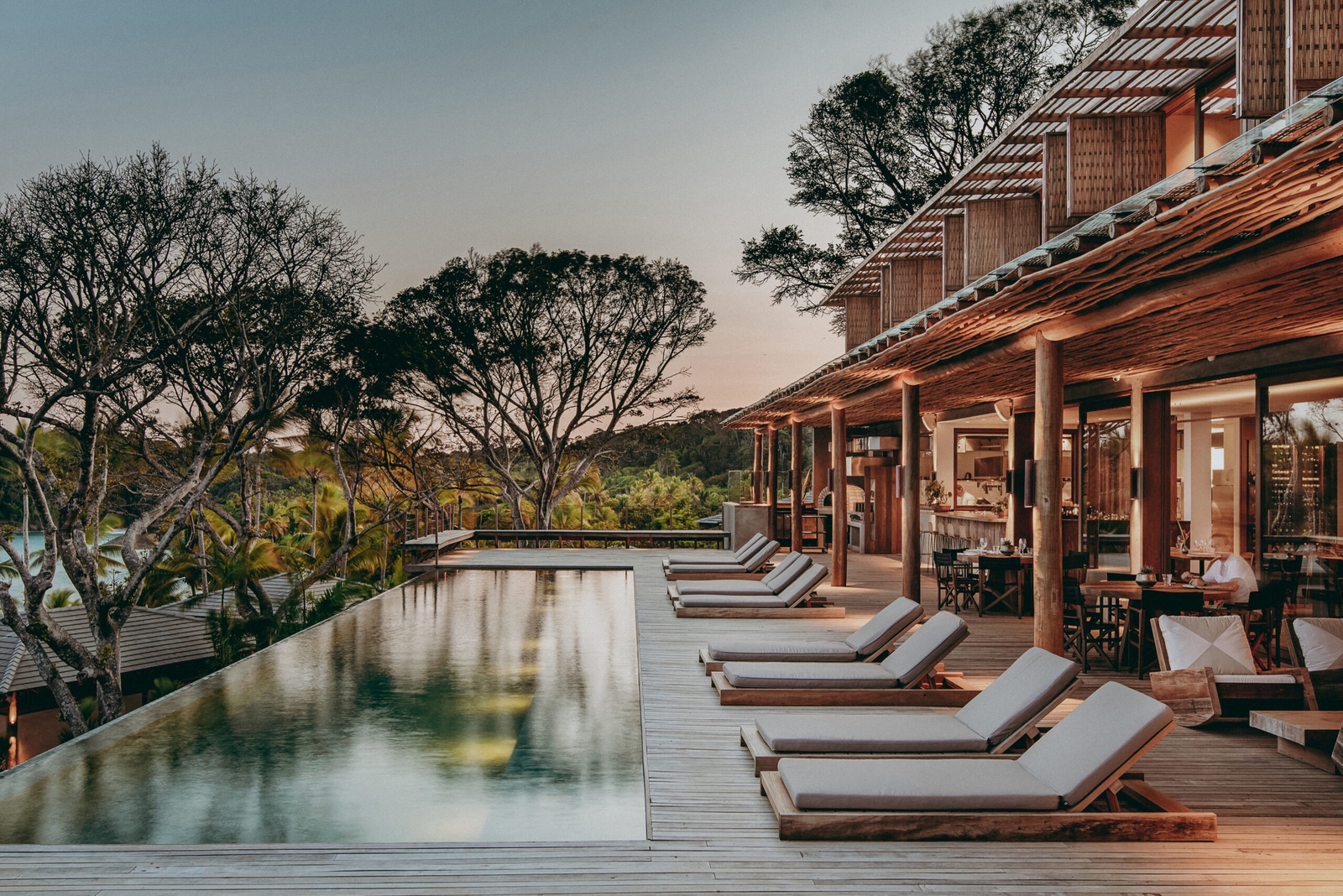 A row of sun loungers with beige cushions overlooking a long pool in front of a two-storey open terrace with trees beyond.