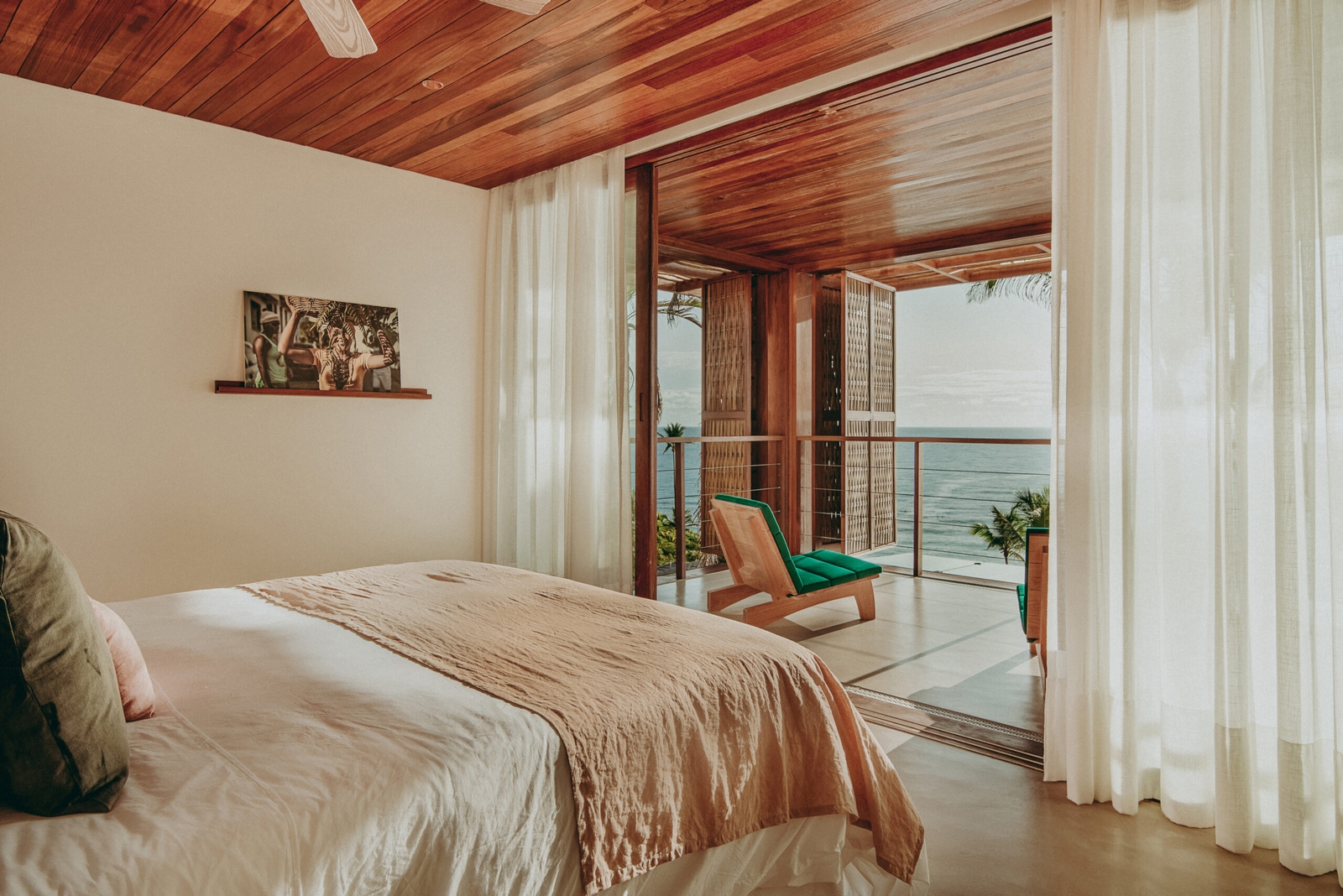Interior view from a double bed looking through an open door framed by white curtains, onto an outdoor terrace with two chairs with blue cushions overlooking a turquoise sea and palm trees.