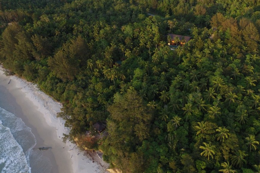 Aerial shot of a thick forest of green trees with a villa set in them and on one side a white sand beach with blue sea.