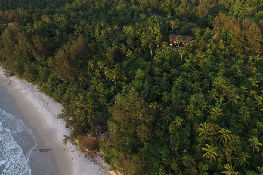 Aerial shot of a thick forest of green trees with a villa set in them and on one side a white sand beach with blue sea.