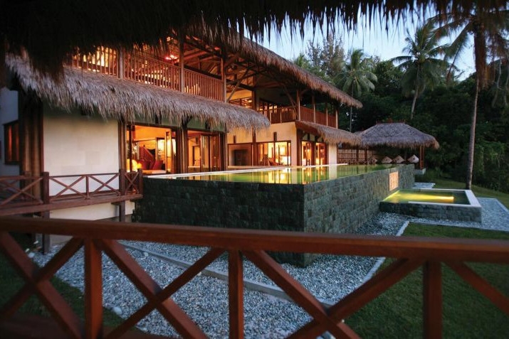 Exterior of a two-storey villa with thatched roof and interior lights on.