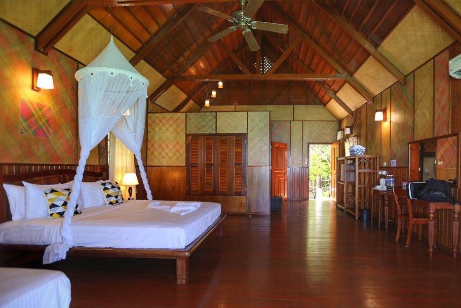 Interior shot of a wooden chalet with yellow walls and a pitched ceiling. Inside is a double bed with white sheets and a mosquito net, and overhead is a ceiling fan.
