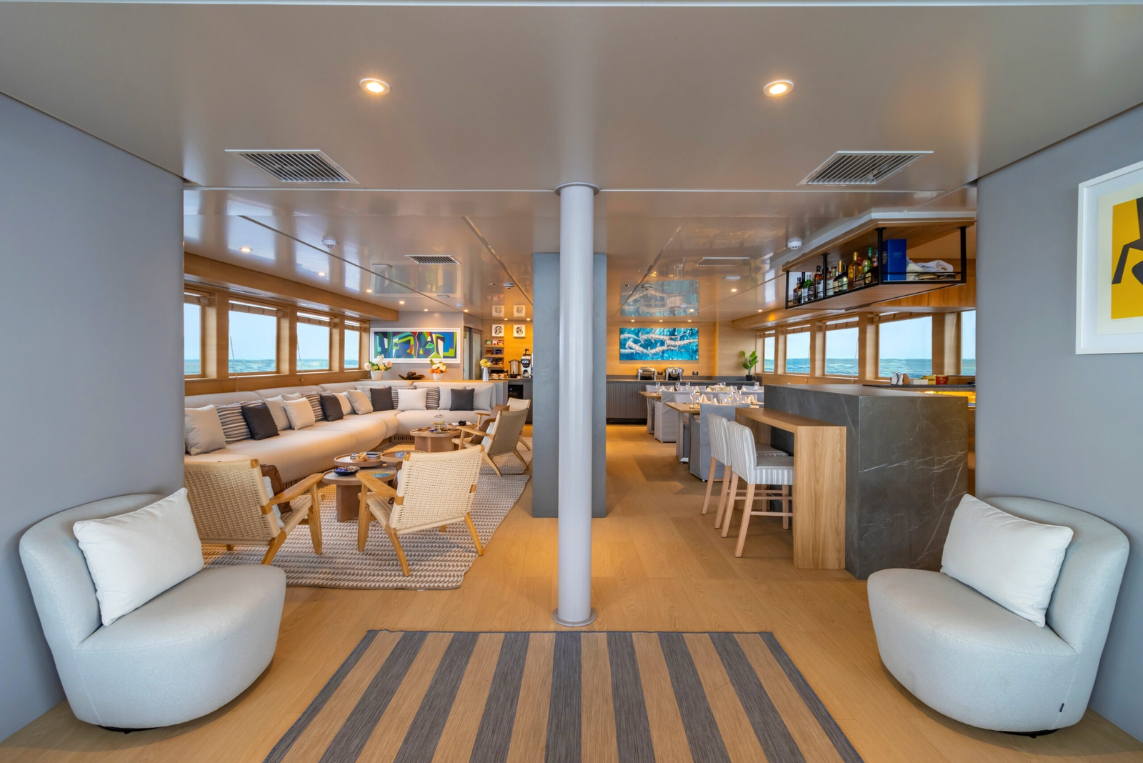 Open seating area with two small plush chairs in the foreground and a pole in the middle of an entryway to a larger seating area with a bar, tables, chairs and banquettes.