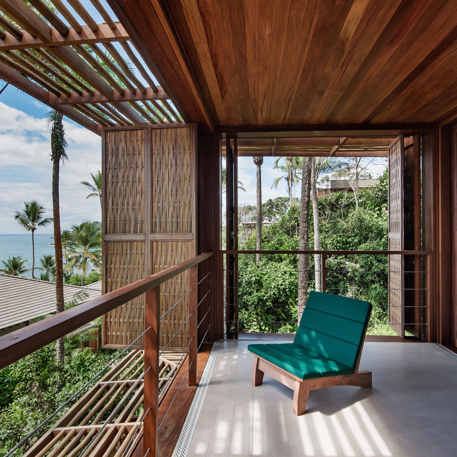 Terrace with red wood roof and thatch sunshade, with one lounge chair with blue cushions overlooking palm jungle and sea.