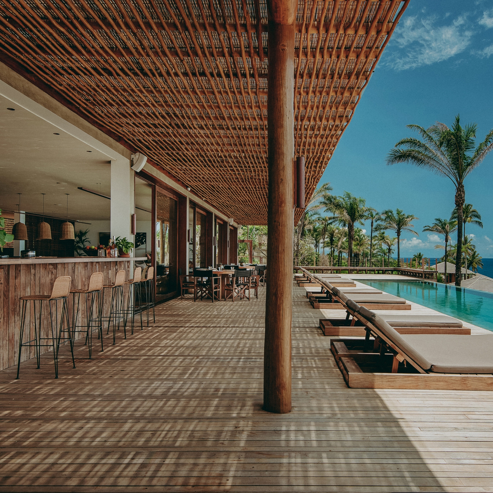 An open-air bar set under a thatch sunshaded terrace with high barstools overlooking a long blue pool, palm trees and blue sky.