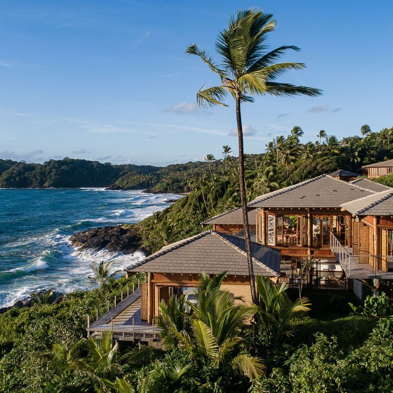 Villa with pitched roof and wrap-around terraces set in thick palm jungle overlooking a rocky coastline with blue ocean and waves crashing.