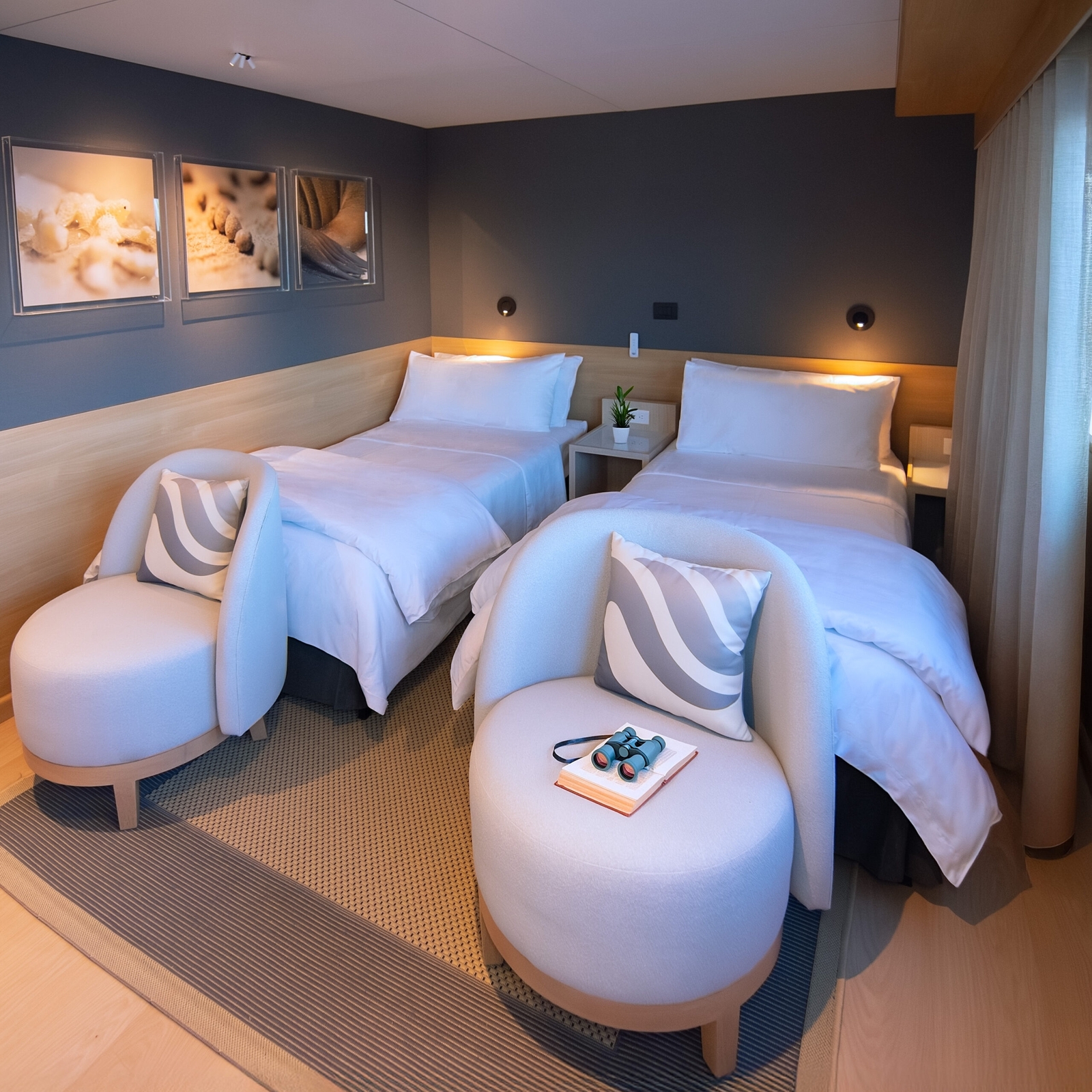 two twin beds with white sheeds and footstools at the base in a carpeted room with grey walls and paintings on one wall.