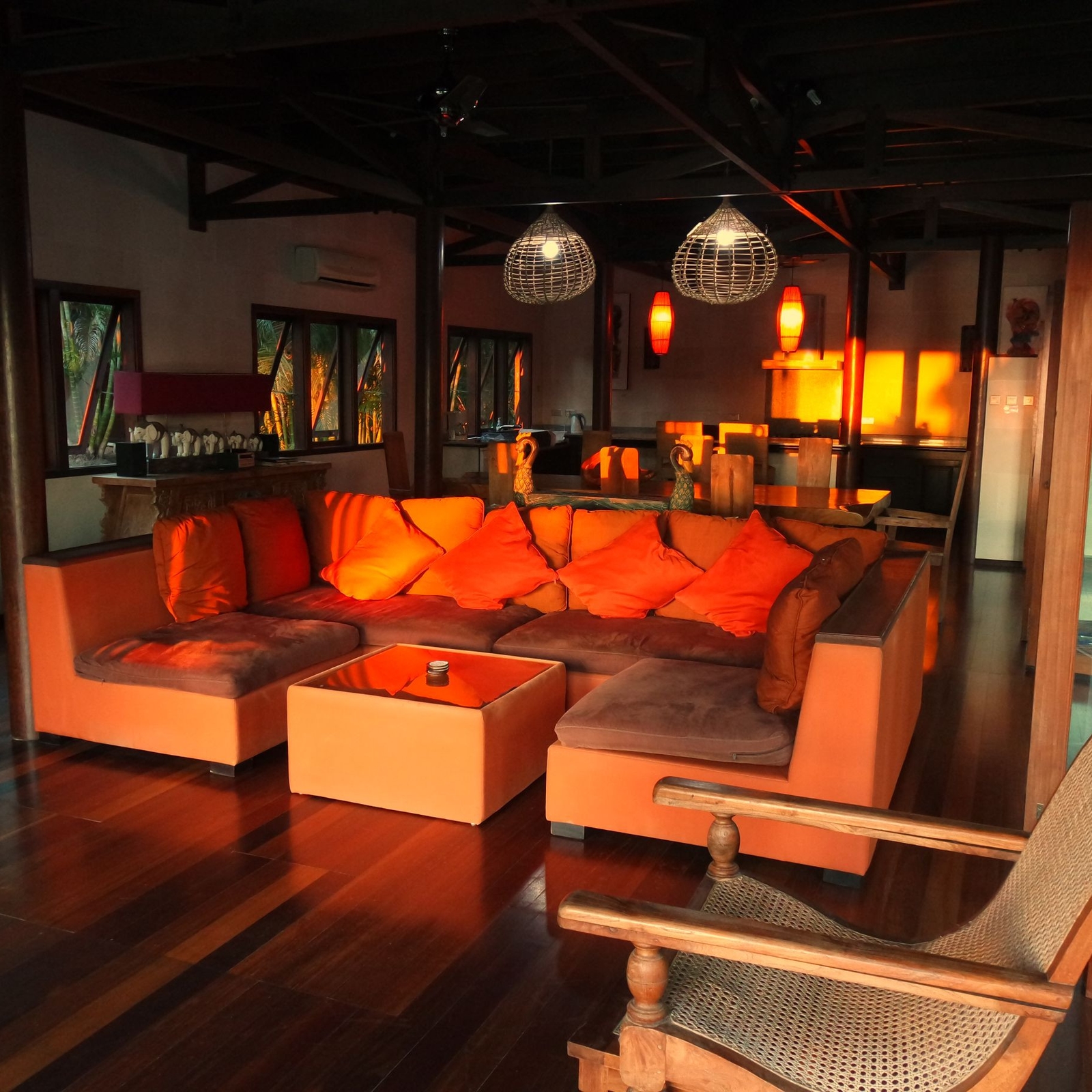 Interior shot of a sunny room with a horseshoe-shaped sofa with orange pillows, a rocking chair, overhead lamps and wood floors.