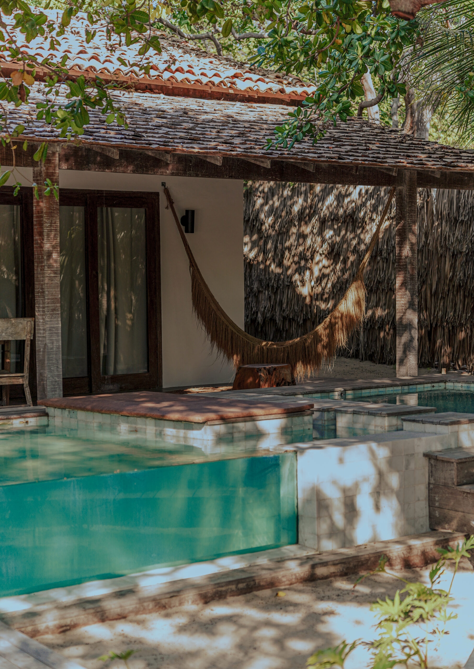 Exterior of a terrace and doorway into a bedroom, with a patio and hanging hammock. In the foreground is a blue pool.
