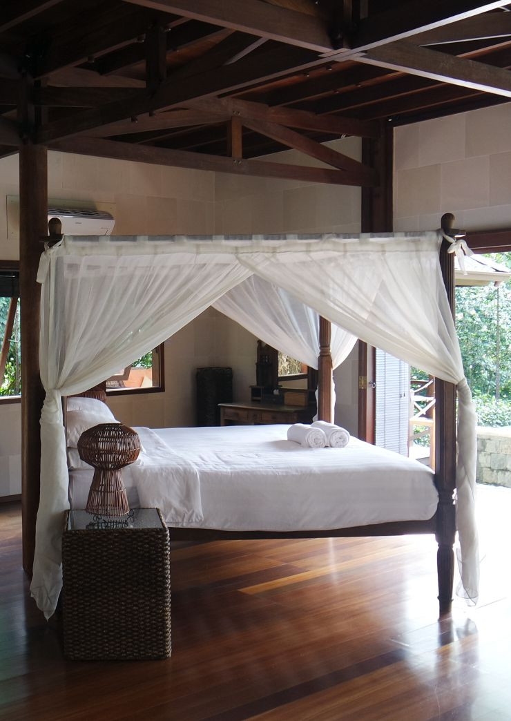 Interior of a bedroom with wood floors and beams, and a four-poster bed with white sheets and mosquito netting. In the background, a door opens onto a terrace.