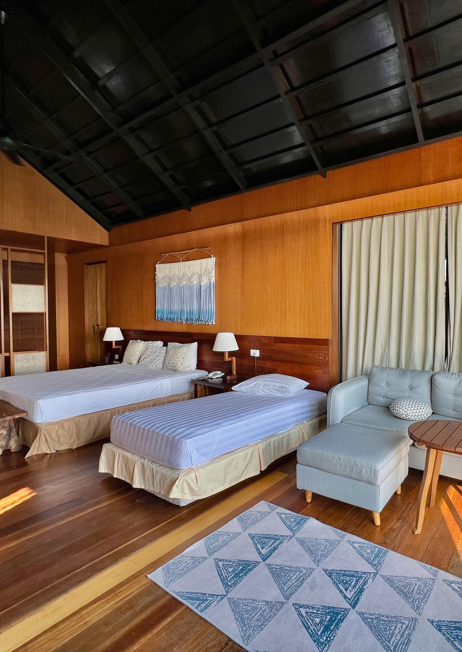 Interior of a bedroom with a double bed and a single bed as well as part of a grey sofa. The walls and floor are hardwood, and there is a grey and blue rug on the floor.