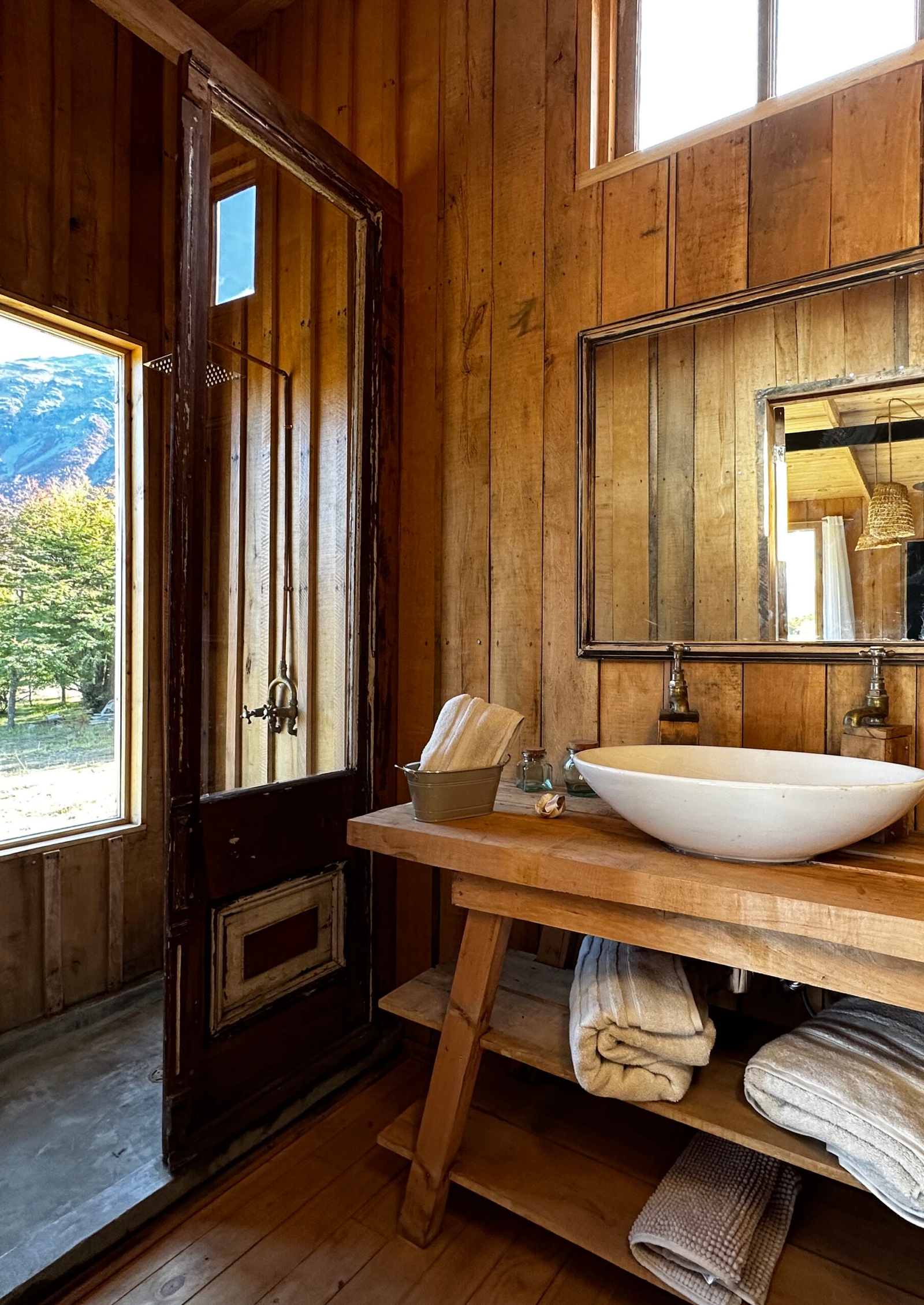 Bathroom with wooden walls, a vessel sink on a rustic timber counter, folded towels and a glass door opening to a shower area with a mountain view.