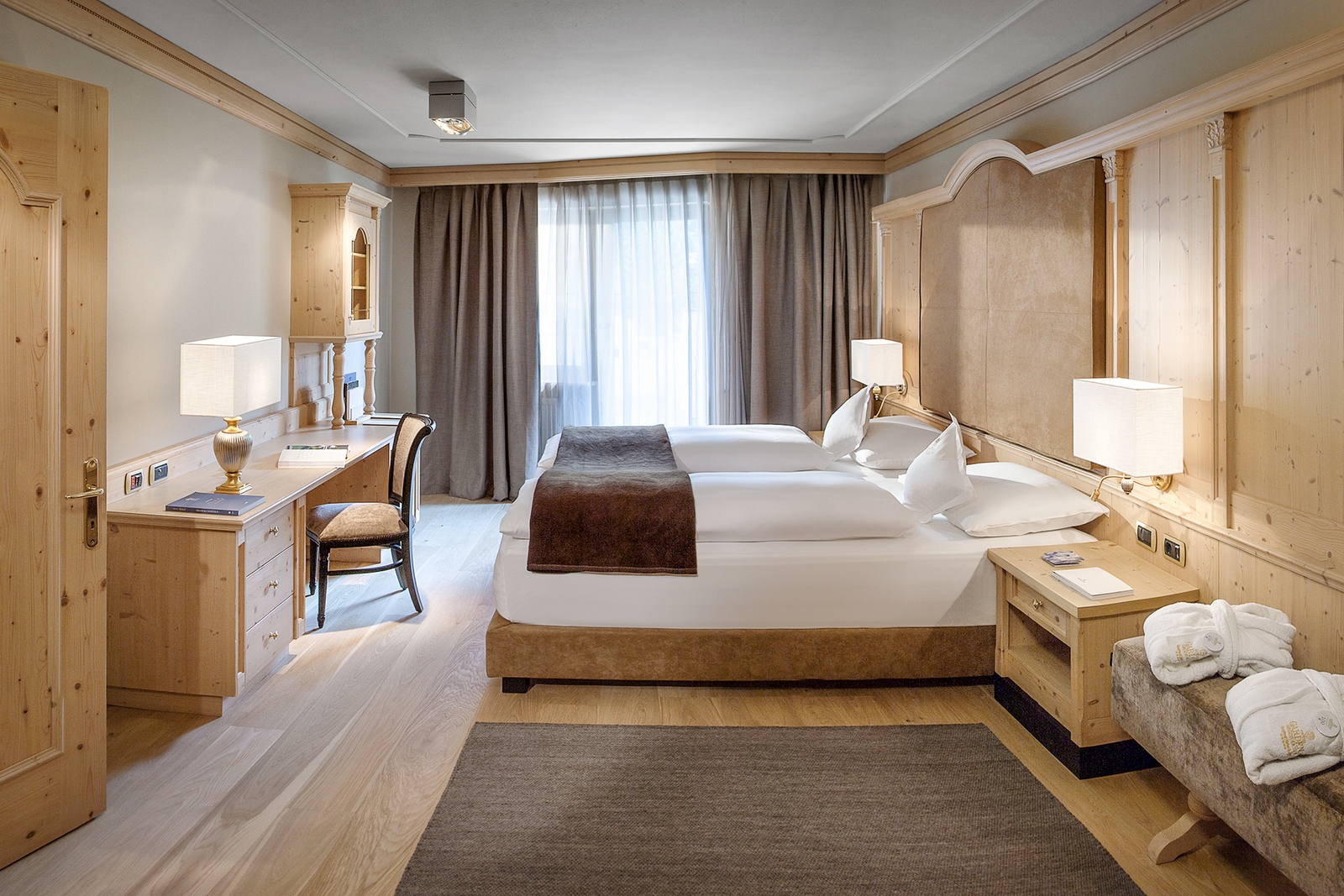 Hotel bedroom with light wood doors, furnishings and floors, with a grey run in the foreground, a lounge chair with two bathrobes, and the bed has a dark brown accent cover.