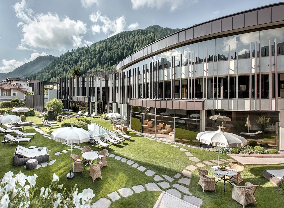Hotel exterior with glass fronted building and green lawn area with white umbrellas and lounge chairs. In the background are mountains with green forests.