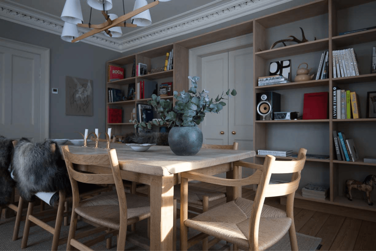 A dining area with a light wood table and chairs, a grey vase of eucalyptus stems and bookshelves in the background