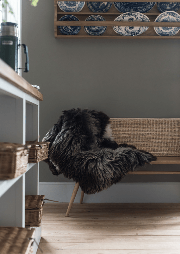 An indoor bench with a grey and white fur blanket draped across it and a shelf of china on the wall above it