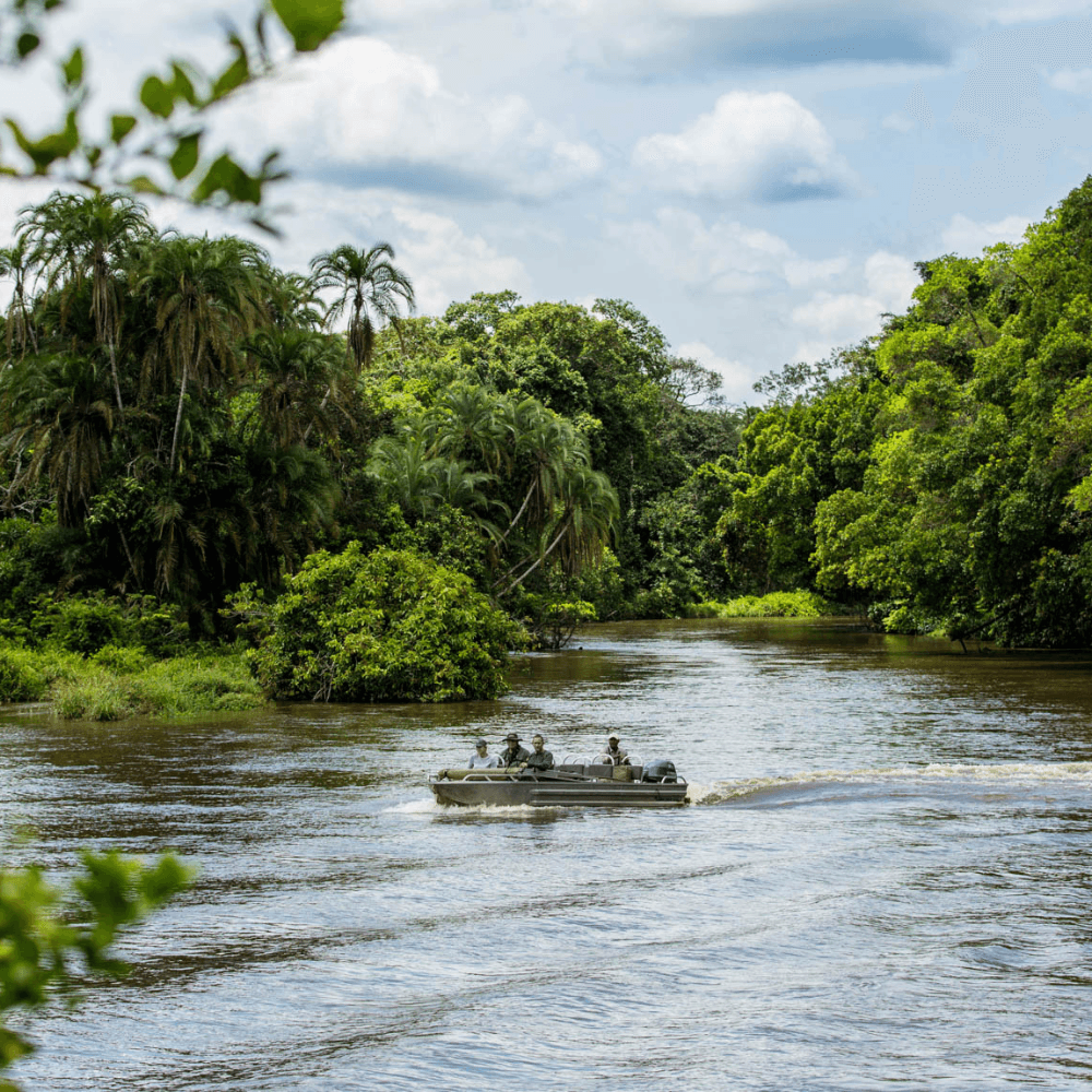 A small boat with four people in it, cruising across a wide river surrounded by green foliage