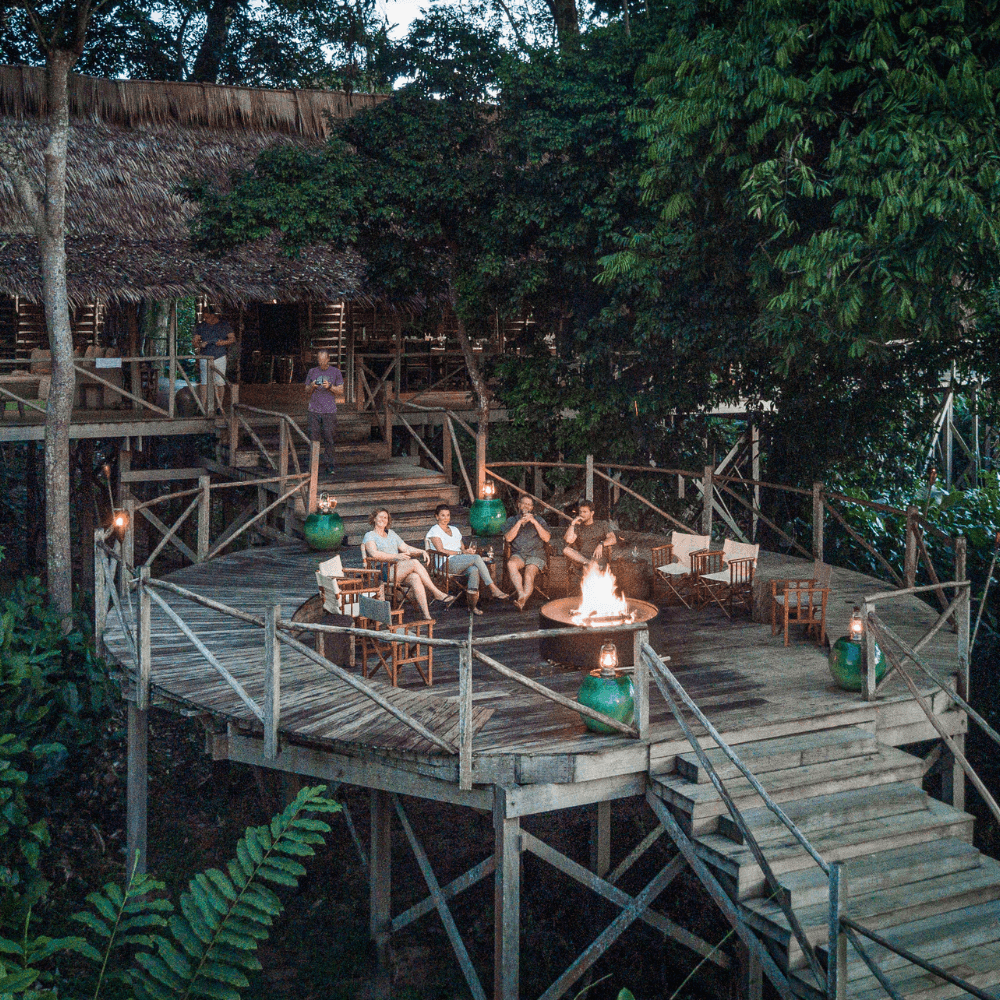 Four smiling people sitting in chairs on a raised round wooden deck with a bonfire