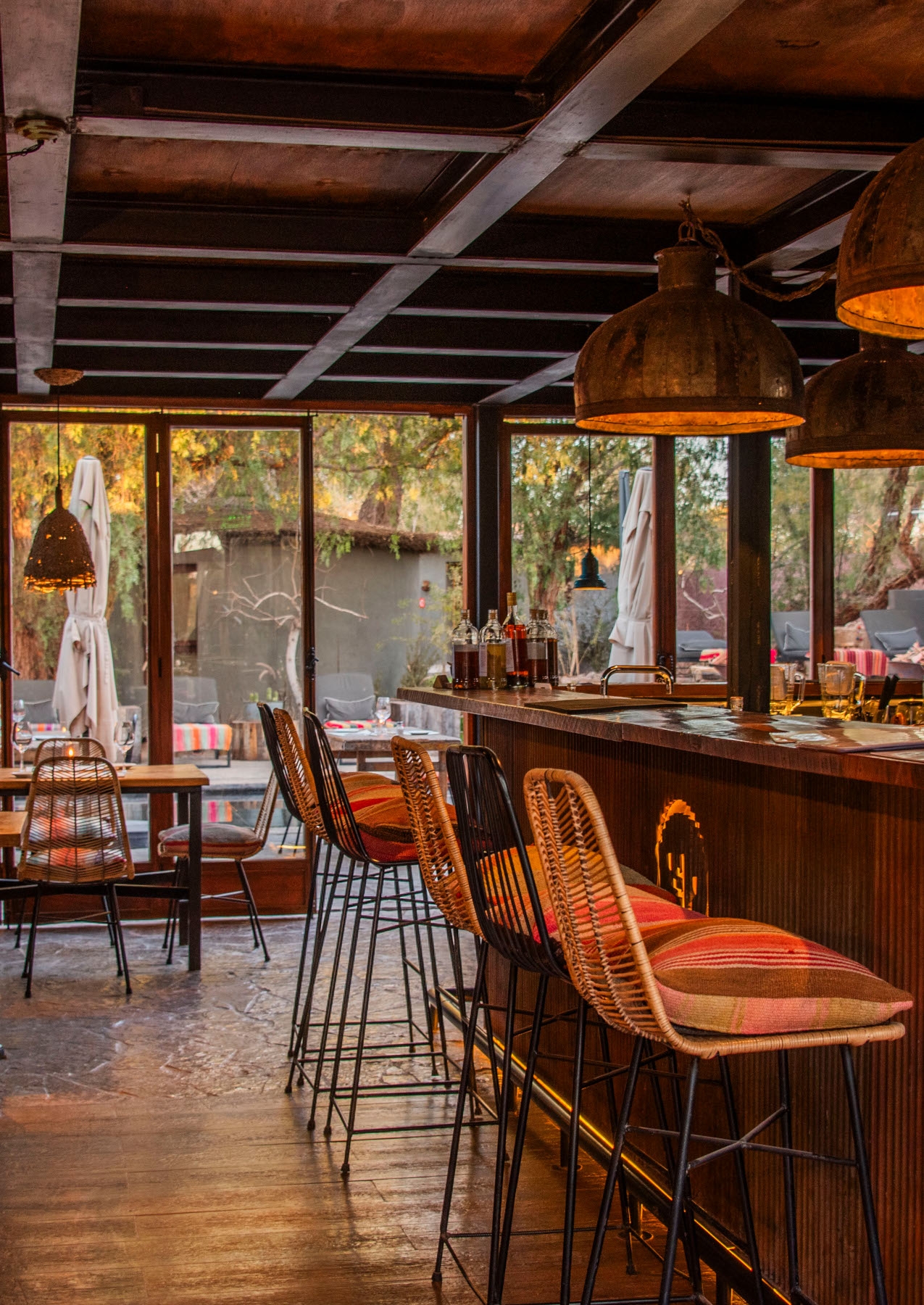 Interior of a bar and restaurant area, with a high bar and high chairs, and in the background are lower tables and an exterior seated patio area visible through glass windows.